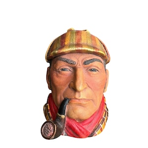 May include: A detailed bust of a man with a pipe, wearing a plaid cap and a red scarf. The sculpture has a weathered appearance, with visible details in the face and clothing. The pipe is black, and the cap features shades of gold, brown, and green.