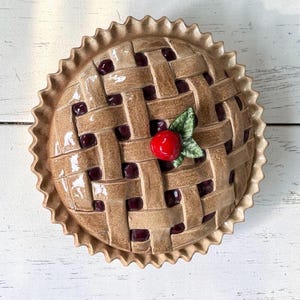 May include: A round ceramic pie dish with a glossy brown lattice crust design. The top features a single bright red cherry with two green leaves centered on the crust. Dark purple filling is visible through the gaps in the woven dough pattern.