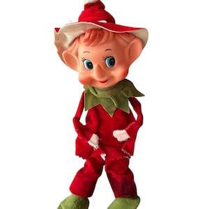 Puede incluir: Muñeca de elfo navideño vintage con un atuendo de terciopelo rojo, cuello verde y zapatos de fieltro verde. El elfo tiene una cara sonriente con ojos azules, mejillas rosadas y un sombrero rojo con borde blanco.