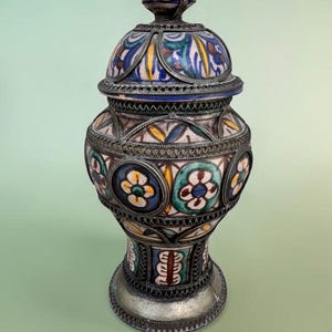 May include: A decorative lidded jar featuring intricate metal filigree overlays on a ceramic body. The ceramic surface displays hand-painted floral motifs in shades of blue, green, yellow, and white, encased in a dark, aged metal framework.
