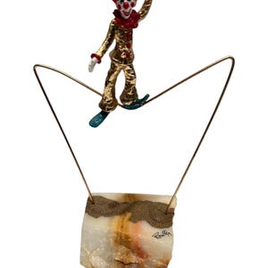 May include: A whimsical sculpture featuring a miniature clown balancing on a wire. The clown is dressed in a gold suit with red accents and a teal hat. The wire is suspended above a rectangular, multi-toned stone base.