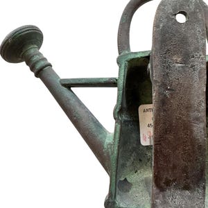 May include: An antique, weathered green metal watering can with a spout, handle, and a rounded top. The can has a label attached. The overall design suggests a vintage or rustic aesthetic, suitable for gardening or decorative purposes.