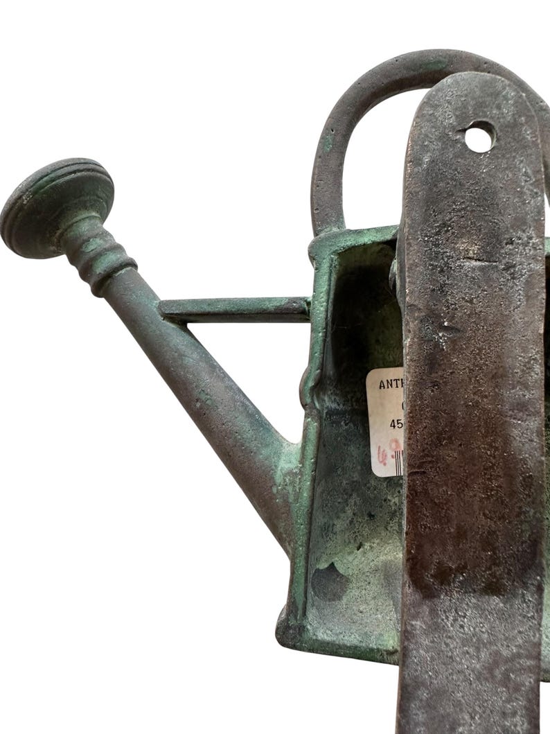 May include: An antique, weathered green metal watering can with a spout, handle, and a rounded top. The can has a label attached. The overall design suggests a vintage or rustic aesthetic, suitable for gardening or decorative purposes.
