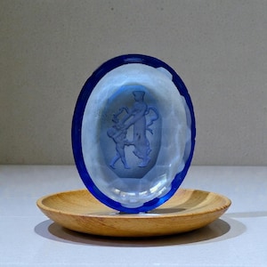 Può includere: Un oggetto di vetro blu a forma ovale con un disegno scolpito di due figure, posto su un piatto di legno. Il vetro ha un bordo sfaccettato e un bordo blu scuro. Le figure sono in stile classico.