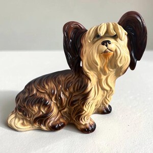 May include: A ceramic figurine of a Yorkshire Terrier dog with long, flowing fur in shades of tan and dark brown. The dog is depicted in a seated position with large, upright ears, a black nose, and detailed sculpted hair texture throughout.