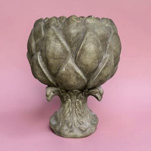 Puede incluir: Una maceta decorativa con forma de alcachofa y acabado en verde apagado desgastado. El cuenco presenta capas superpuestas que recuerdan a pétalos, mientras que la base está esculpida con detalles de hojas, creando un aspecto orgánico y texturizado.