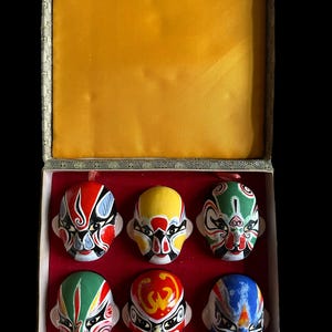 Puede incluir: Un conjunto de seis pequeñas máscaras de ópera china coloridas, presentadas en una caja rectangular. Las máscaras presentan diseños pintados intrincados en rojo, amarillo, verde y azul, con detalles en blanco y negro. La caja tiene un forro de satén dorado.
