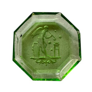 Può includere: Un posacenere ottagonale in vetro verde. Il posacenere presenta un rilievo dettagliato di una figura alata che suona un flauto, con elementi architettonici. Il vetro ha una leggera consistenza ed è trasparente.