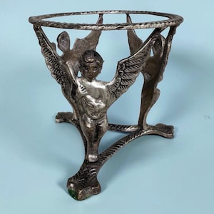 Puede incluir: Un soporte de metal plateado con tres figuras de querubines con alas extendidas. Los querubines están conectados por un anillo superior circular y se extienden hacia abajo para formar una base de trípode. El metal tiene un acabado texturizado.