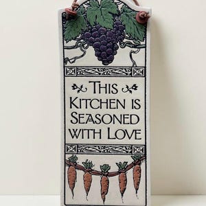 May include: A rectangular ceramic wall plaque with a cream background, featuring a purple grape cluster at the top and a row of orange carrots at the bottom. The text reads, "THIS KITCHEN IS SEASONED WITH LOVE." It includes a brown leather hanging cord.