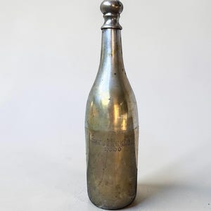 May include: A silver-toned bottle with a rounded top and a spherical stopper. The bottle has a label that reads "Millennium 2000". The bottle is made of metal and has a vintage appearance.