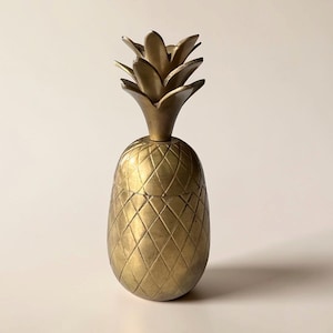 Może przedstawiać: Złoty, dekoracyjny przedmiot w kształcie ananasa. Ananas ma teksturowany, diamentowy korpus i liściasty wierzch. Obiekt jest prawdopodobnie wykonany z metalu i przeznaczony do celów dekoracyjnych.