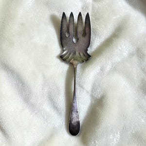 May include: A silver-toned metal serving fork featuring a wide, flat head with four short, pointed tines and a decorative scalloped base. The handle is slender and tapers toward a rounded end, with visible tarnishing across the surface.