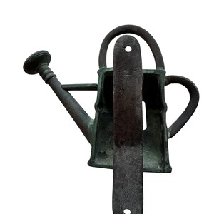 May include: A weathered, bronze-colored watering can with a long spout and a handle. A dark, rectangular metal plate is positioned in front of the watering can. The watering can has a classic design, suitable for gardening.