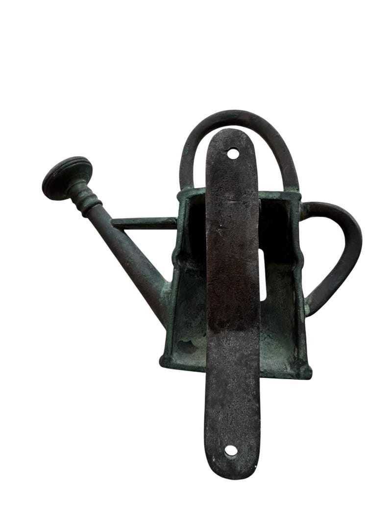 May include: A weathered, bronze-colored watering can with a long spout and a handle. A dark, rectangular metal plate is positioned in front of the watering can. The watering can has a classic design, suitable for gardening.