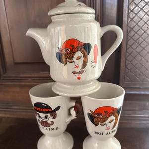 May include: A vintage ceramic tea set comprising a teapot and two pedestal cups. The teapot and one cup depict a woman in a red hat, while the other cup shows a man in a hat. The cups have the text "Je t'aime" and "Moi aussi".