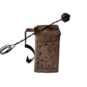 May include: An antique metal container, rectangular with a hinged lid, showing signs of age and wear. It features a wire handle and a small, spherical object at the end. The metal has a dark, textured finish with visible rivets.