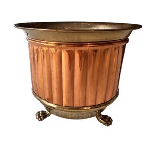 Puede incluir: Una maceta de color cobre con un diseño acanalado y un borde de latón. La maceta tiene una base de latón con tres patas de garra. El borde superior también es de latón. Es un artículo decorativo.