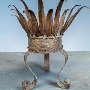 Può includere: Un supporto decorativo in metallo con una base cilindrica centrale dalla texture martellata e un bordo intrecciato. La parte superiore è decorata con punte metalliche a forma di foglia rivolte verso l'alto e poggia su tre gambe ricurve.