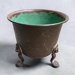 May include: A round, deep metal planter with a dark brown exterior and a bright green interior patina. The vessel features a wide, flared rim and is supported by three distinct, sculpted feet that resemble small, stylized animal figures.