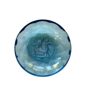 Può includere: Una ciotola di vetro blu chiaro con un design in rilievo dettagliato al centro. Il disegno raffigura una figura inginocchiata con una figura più piccola in piedi nelle vicinanze. La ciotola ha un bordo smerlato ed è su sfondo bianco.