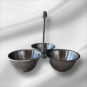 May include: A stainless steel serving dish with three bowls and a central handle. The bowls are arranged in a triangular pattern, perfect for serving dips, sauces, or snacks. The metal has a brushed finish, giving it a modern look.