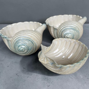 Peut inclure: Trois bols en céramique en forme de coquillages avec un intérieur crème brillant et un extérieur texturé présentant un motif en spirale dans des tons de bleu et de blanc. Chaque bol a un bord festonné et une forme de coquillage distincte.