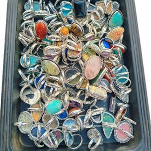 May include: A collection of assorted rings with various colorful gemstones set in silver-toned metal bands, all contained within a black rectangular tray. The rings feature diverse shapes including oval, rectangular, and teardrop cabochons.