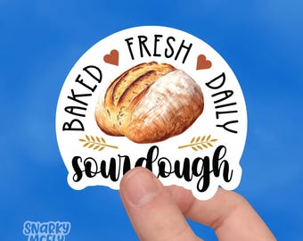 May include: White round sticker with the text "Baked Fresh Daily Sourdough" in black lettering. A brown heart is on either side of the text "Fresh". A golden wheat stalk is on either side of the text "Sourdough". A loaf of sourdough bread is in the center of the sticker.