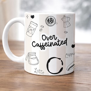 Peut inclure: Un mug en céramique blanche avec des rayures verticales beiges et des illustrations au trait noir représentant des articles de café, dont une cafetière italienne, un latte et des grains de café. Le texte Over Caffeinated est écrit au centre.