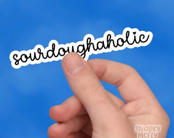 May include: White sticker with black text that reads "sourdoughaholic" with a small heart symbol. The sticker is being held by a hand against a blue background.