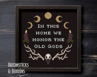 Puede incluir: Un bordado de punto de cruz enmarcado en negro con el texto "In this home we honor the old gods" rodeado de un diseño de luna creciente y velas. El texto "Broomsticks & Bobbins" está en la esquina inferior izquierda.