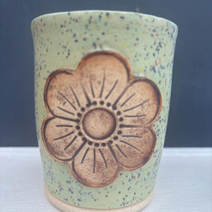 Könnte beinhalten: Handgefertigte Keramiktasse mit hellgrüner, gesprenkelter Glasur. Auf der Vorderseite ist ein großes, braunes Blumenmuster geprägt. Die Tasse hat eine abgerundete Form und einen natürlichen, unglasierten Boden.