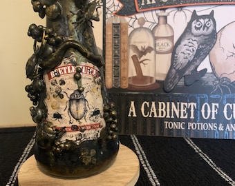 Pode incluir: Uma garrafa de vidro preto com um rótulo dourado que diz "Beetle Juice" e apresenta uma ilustração de um besouro. A garrafa é decorada com detalhes dourados e pretos e tem uma base de madeira.