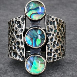 May include: A silver ring featuring three iridescent abalone shell insets. The ring has a textured band with a hammered metal design. The abalone shells display green, blue, and purple hues.