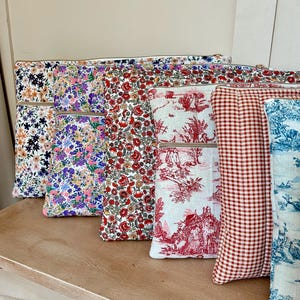 Könnte beinhalten: Eine Auswahl an Reißverschlusstaschen mit verschiedenen floralen und gemusterten Stoffen. Designs umfassen marineblaue Blumen, lila Blumen, rote Blumen, rotes Toile de Jouy, rotes Vichy-Karo und blaues Toile de Jouy. Die Taschen liegen auf einer hellen Holzoberfläche.