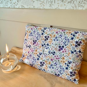 Peut inclure: Une pochette blanche à fermeture éclair avec un motif floral dans les tons rose, violet, bleu et jaune. La pochette est posée sur une surface en bois.