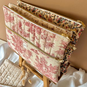Peut inclure: Trois pochettes zippées matelassées avec un motif toile rouge et blanc. Les pochettes sont empilées les unes sur les autres et ont des fermetures éclair dorées.