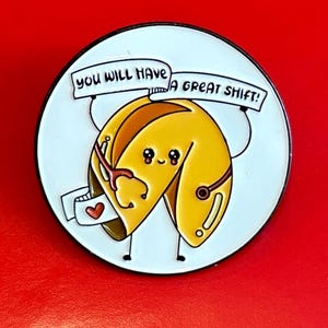 May include: A white enamel pin with a cartoon fortune cookie holding a banner that says "You will have a great shift!" The fortune cookie is yellow with a red heart on the fortune paper.