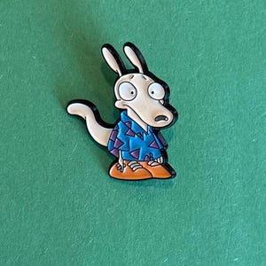 May include: A cartoon character pin featuring a light tan kangaroo-like creature with large eyes and a surprised expression. The character wears a blue shirt with a geometric pattern and orange pants. The pin has a black outline and is set against a green background.