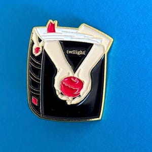 May include: Gold-colored enamel pin featuring a black book with a red apple in the center. The book has the word "twilight" written on the cover. The apple is being held by two white hands.