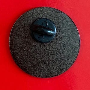 May include: The back of a round, black pin with a metal pin back.