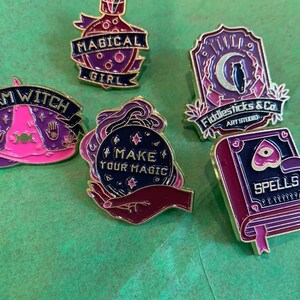 May include: Five enamel pins with a witch theme. The pins feature a purple potion bottle with the text "Magical Girl", a purple book with the text "Spells", a purple crystal ball with the text "Make Your Magic", a purple owl with the text "Fiddlesticks & Co. Art Studio", and a pink witch hat with the text "Witch".