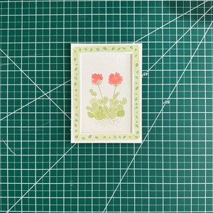 May include: A rectangular white card featuring a watercolor illustration of two coral-pink geranium flowers with light green leaves and visible roots, framed by a light green border with small dark green dots, measuring 3 inches by 5 inches.