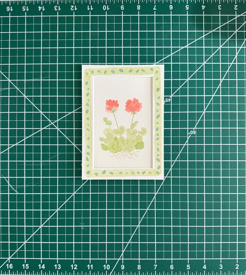 May include: A rectangular white card featuring a watercolor illustration of two coral-pink geranium flowers with light green leaves and visible roots, framed by a light green border with small dark green dots, measuring 3 inches by 5 inches.