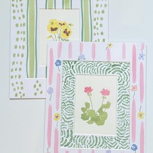 May include: Two rectangular art prints featuring botanical illustrations. The top print displays yellow pansies with a green striped border and a white frame with green dashes. The bottom print shows pink flowers with a green wavy border and pink stripes.