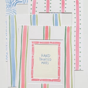 May include: A collection of three rectangular paper picture frame mats featuring hand-painted designs. One mat has blue wavy lines, another has pink stripes with green dots, and the front mat has pink and green stripes with the text HAND PAINTED MATS.