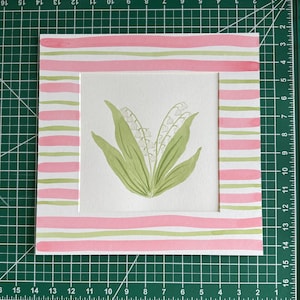 May include: A square watercolor painting featuring a central illustration of lily of the valley flowers with green leaves and white blossoms. The artwork is framed by a border of horizontal pink and light green stripes on a white background.