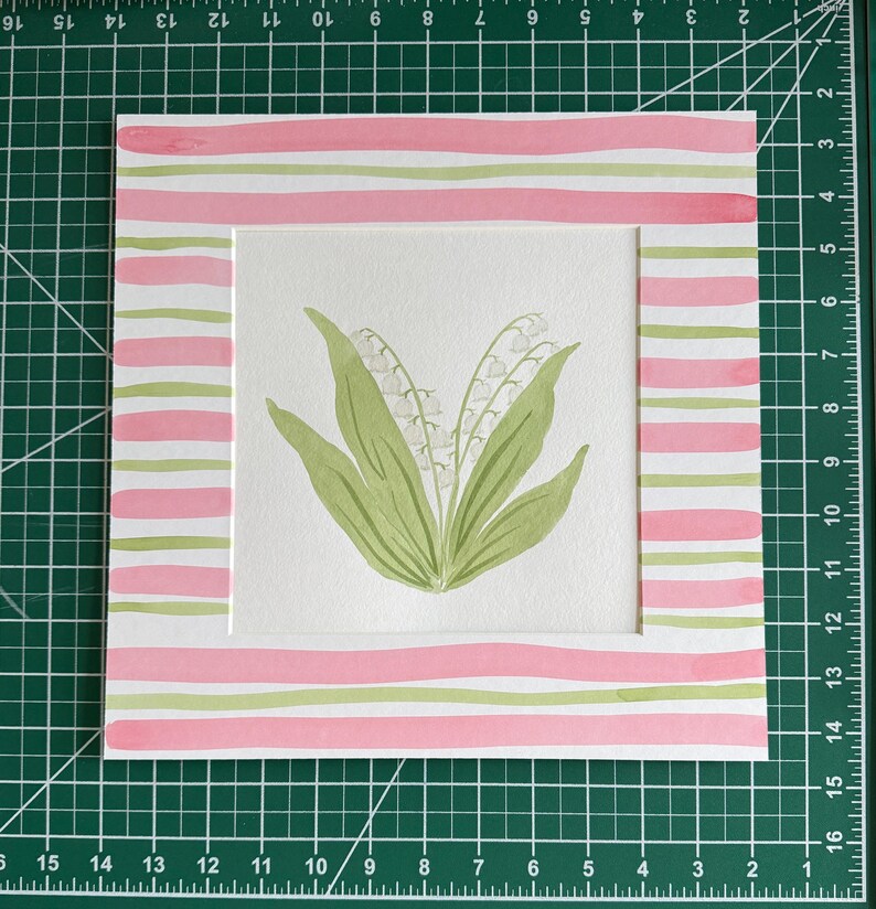 May include: A square watercolor painting featuring a central illustration of lily of the valley flowers with green leaves and white blossoms. The artwork is framed by a border of horizontal pink and light green stripes on a white background.