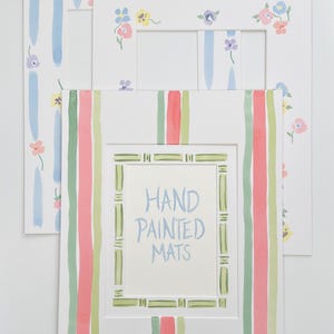 May include: Three white mat boards with hand-painted borders. The top mat has a blue and white striped border with small flowers. The middle mat has a floral border with yellow, blue, and pink flowers. The bottom mat has a green and pink striped border. The inner most mat has a green border with the text "HAND PAINTED MATS".